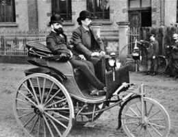 Two men riding in a tricycle automobile in the 19th century